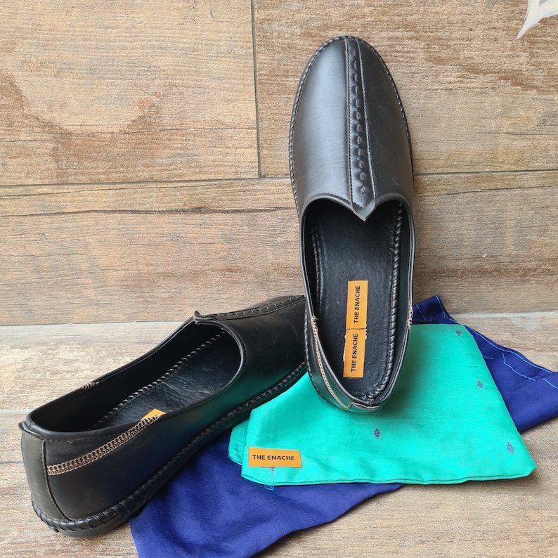 Op de afbeelding: Zwarte leren instappers met decoratieve stiksels en een ronde neus. De schoenen worden getoond met een turquoise en marineblauwe stof. De tekst "THE ENACHE" is zichtbaar op de schoenen en de stof.
