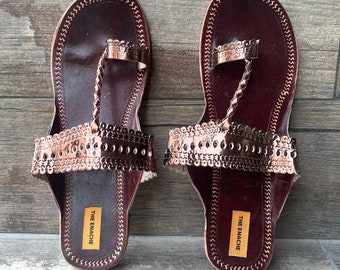 May include: A pair of brown leather sandals with a gold-colored trim and a toe ring. The sandals have a brand label that reads "THE ENACHE".