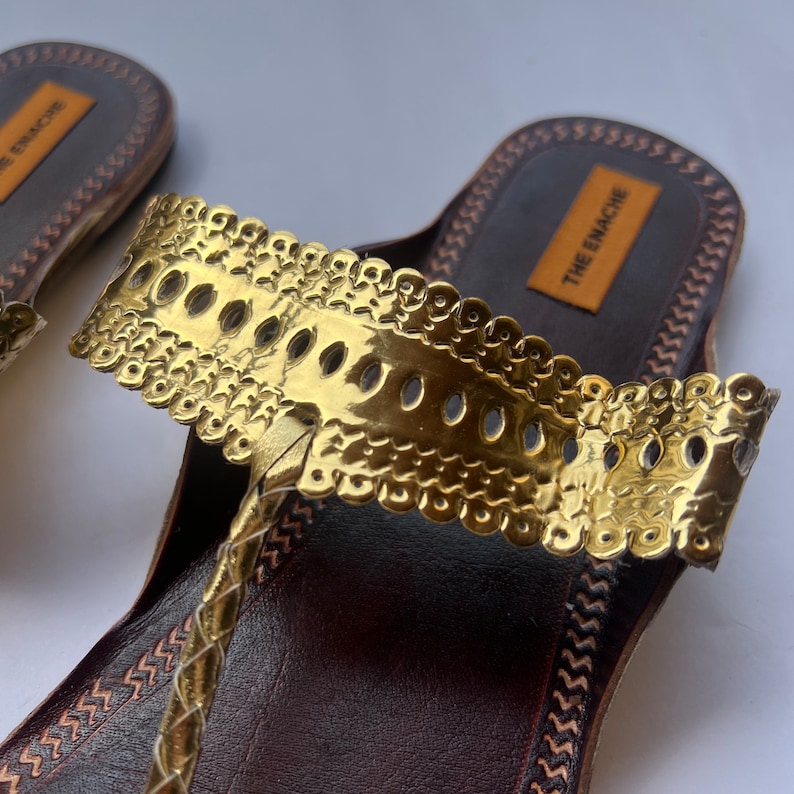 May include: Gold leather sandals with a decorative strap featuring a gold embossed design and the text "THE ENACHE" on the sole.
