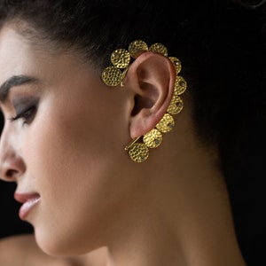 Peut inclure: Une boucle d'oreille dorée en forme de courbe avec plusieurs disques ronds texturés. Chaque disque a une petite pierre précieuse transparente au centre.