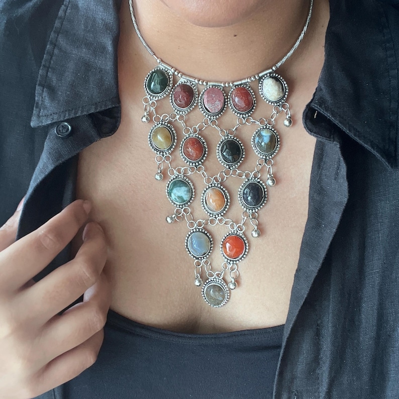 May include: A silver-toned necklace featuring a cascade of oval-shaped gemstones in various colors, including green, red, and orange. The gemstones are set in a decorative silver frame and connected by delicate chains, with small dangling accents.