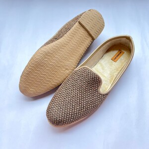 Op de afbeelding: Een paar gouden geweven slippers met een bruine rubberen zool. De slippers hebben een label met de tekst "The Encore".
