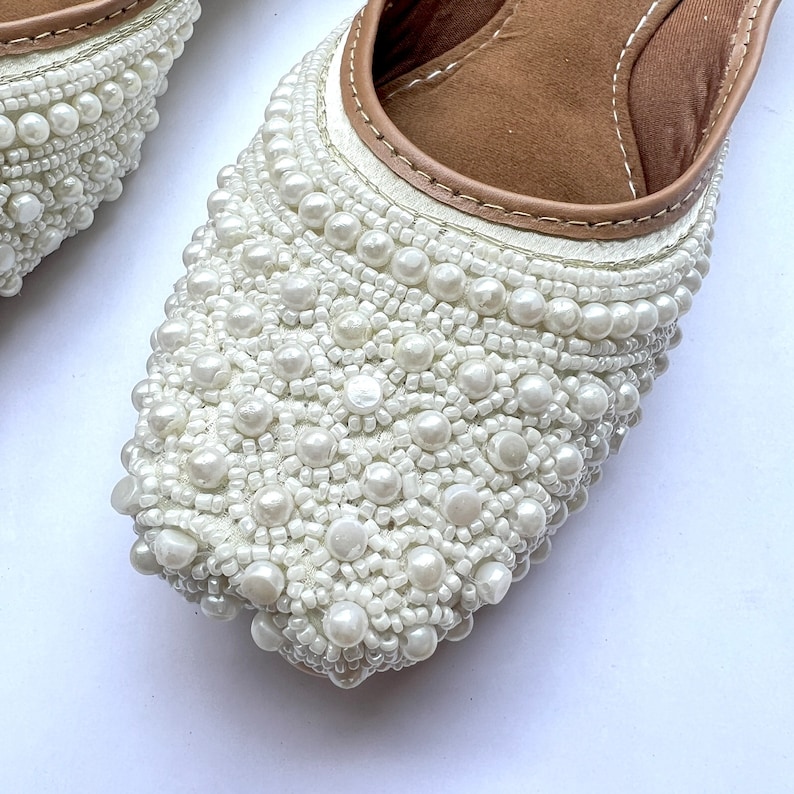 May include: White flat shoes with a rounded toe and a white pearl embellishment covering the entire shoe.