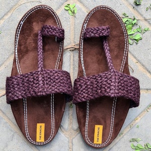 May include: A pair of brown leather sandals with a purple jute strap and a white stitched border. The sandals have a toe loop and the brand name "THE ENACHE" is printed on the bottom of each sandal.