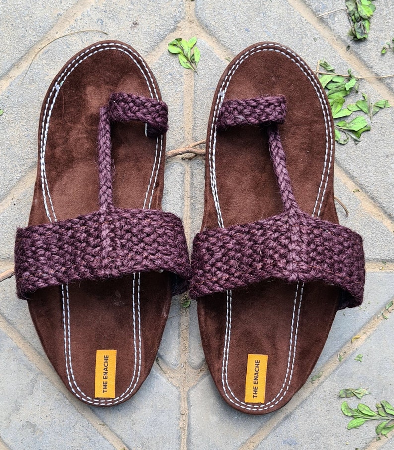May include: A pair of brown leather sandals with a purple jute strap and a white stitched border. The sandals have a toe loop and the brand name "THE ENACHE" is printed on the bottom of each sandal.
