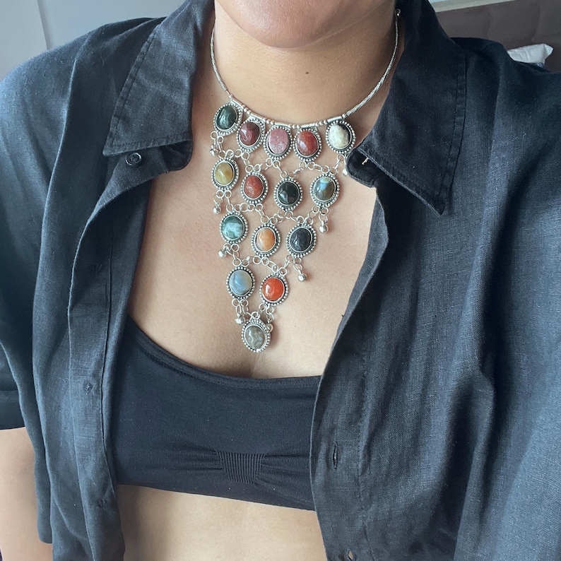 May include: A silver-toned necklace with a cascade of colorful, oval-shaped gemstones. The necklace features a variety of colors, including green, red, orange, and black, set in ornate silver frames. Small silver dangles hang from the bottom of the necklace.