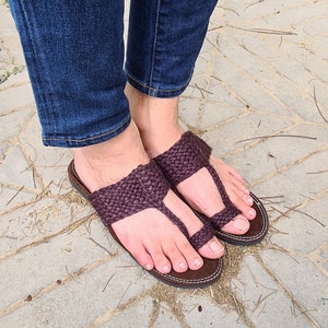 May include: A pair of brown woven sandals with a thong style strap. The sandals are worn on a person's feet with denim jeans.