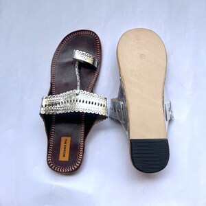 May include: A pair of silver leather sandals with a toe ring and a brown leather sole. The sandals have a decorative silver trim and the text "THE ENACHE" is printed on the sole.