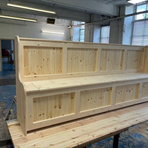 May include: A long wooden bench with a high back and storage compartments underneath. The bench is made of natural wood and has a simple, rustic design.