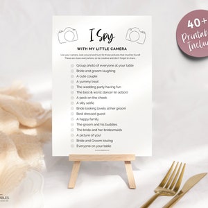 May include: A printable I Spy game with a camera theme. The game features a list of items to find at a wedding, such as a group photo, the bride and groom laughing, a cute couple, a yummy treat, and more.