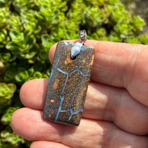Op de afbeelding: Een rechthoekige boulder-opaal hanger met een donkerbruine matrix en natuurlijke, iriserende blauwe en groene aders die door de steen lopen. De hanger is voorzien van een gepolijst zilverkleurig metalen oogje voor bevestiging.