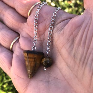 Puede incluir: Un péndulo con un colgante de piedra ojo de tigre triangular y facetado unido a una cadena de metal plateado. Una pequeña cuenta redonda de ojo de tigre también está unida a la cadena, que presenta una serie de eslabones ovalados interconectados.