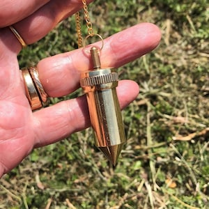 Può includere: Un pendolo color oro con una punta, sospeso a una delicata catena d'oro. Il pendolo ha un corpo cilindrico con una parte superiore strutturata. Lo sfondo è erba verde sfocata.