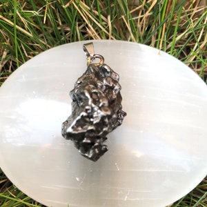 May include: A dark, textured meteorite pendant with a silver-coloured bail. The pendant is irregularly shaped and appears to be made of a metallic material. The pendant is displayed on a white, oval-shaped surface, with green grass in the background.