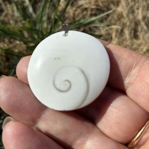 以下が含まれることがあります： 薄い茶色の自然な渦巻き模様が特徴の円形の白いペンダント。ペンダントは滑らかで光沢のある表面を持ち、上部には取り付け用の小さな金属製のベイルが付いており、貝殻のような独特の有機的な質感が際立っています。