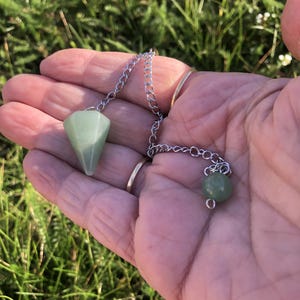Puede incluir: Un colgante de piedra de color verde claro, facetado y en forma de cono, unido a una cadena de metal plateado. La cadena presenta una pequeña cuenta de piedra redonda de color verde claro en el extremo. El colgante tiene una superficie pulida.