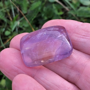 Peut inclure: Une pierre précieuse d'améthyste rectangulaire polie avec une teinte violet clair translucide. La pierre présente des bords lisses et arrondis ainsi qu'une surface plane, révélant des inclusions internes naturelles et des variations de couleur.