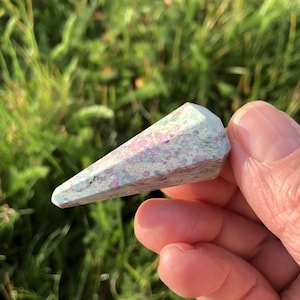 May include: A gemstone crystal point with a light blue and pink speckled pattern. The crystal is held in a hand against a blurred green background of grass. The crystal is a tapered shape, with a flat top and a pointed end.