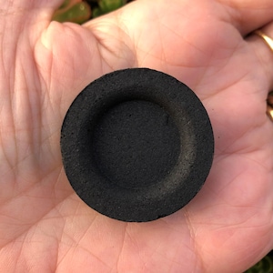 Pode incluir: Um pequeno disco circular de carvão preto com um centro rebaixado. O objeto tem uma textura mate e porosa e uma cor escura uniforme em toda a sua superfície. Tem a forma de uma pastilha redonda e plana com uma indentação central.