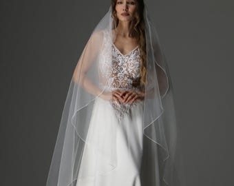 May include: A white wedding dress with a V-neck bodice featuring intricate lace detailing. The dress has a flowing skirt and is paired with a sheer, long veil. The model is wearing the dress in a studio setting.