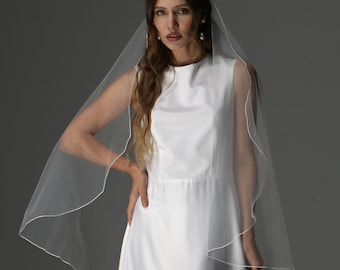 May include: A sleeveless white wedding dress with a flowing skirt and a sheer veil with a white trim. The veil drapes over the shoulders. The model is wearing pearl earrings.