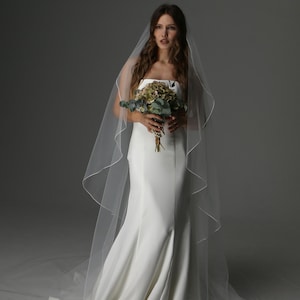 May include: A bride in a white strapless wedding dress with a mermaid silhouette. She wears a long, sheer veil and holds a bouquet of green and brown flowers. The background is a neutral gray.