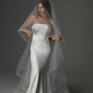 May include: A strapless white wedding dress with a mermaid silhouette. The bride is wearing a sheer, sparkling veil that cascades down the back. The veil is held in place by the bride's hands.