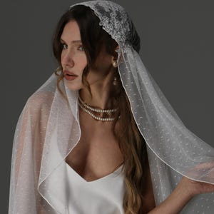 Può includere: Un velo da sposa bianco con un delicato motivo a pois e un copricapo in pizzo. Il velo è drappeggiato su un abito bianco e la modella indossa una collana e orecchini di perle. L'estetica generale è elegante e romantica.