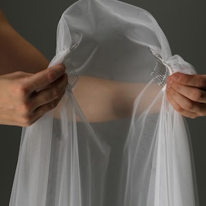 May include: A sheer white bridal veil with a comb attachment. The veil is made of a delicate, lightweight fabric and is held up by two silver combs. The veil is being held up by two hands against a gray background.