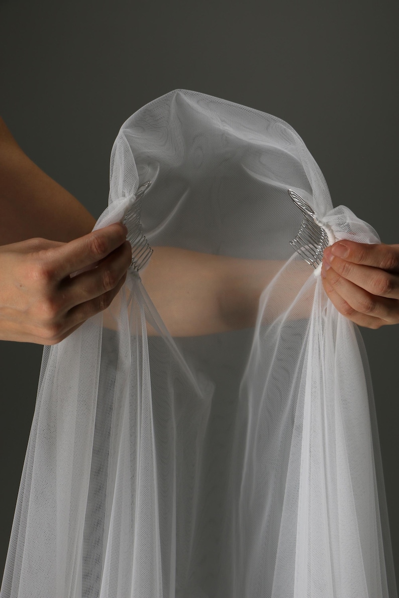 May include: A sheer white bridal veil with a comb attachment. The veil is made of a delicate, lightweight fabric and is held up by two silver combs. The veil is being held up by two hands against a gray background.