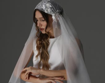 May include: A white wedding veil with a head covering, adorned with delicate lace and sparkling embellishments. The veil cascades down, creating a soft, ethereal look. The bride is wearing a white dress.