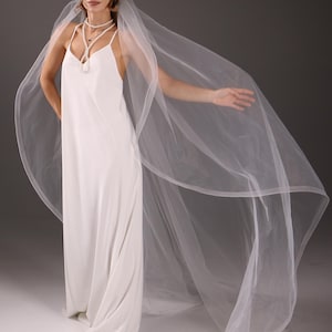 May include: A white wedding dress with a flowing veil. The dress has a simple, elegant design with spaghetti straps. The veil is sheer and extends beyond the dress, creating a graceful, ethereal look. A pearl necklace adds a touch of sophistication.