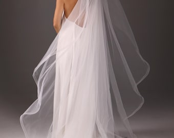 May include: A white wedding dress with a long train and a sheer veil. The dress has a low back and spaghetti straps. The veil is long and flowing, and the model is turned away from the camera.