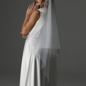 May include: A white bridal veil with a delicate, embellished headpiece. The veil is layered and falls gracefully. The bride is wearing a sleeveless white dress. The image is set against a neutral gray backdrop, highlighting the elegant details of the bridal wear.