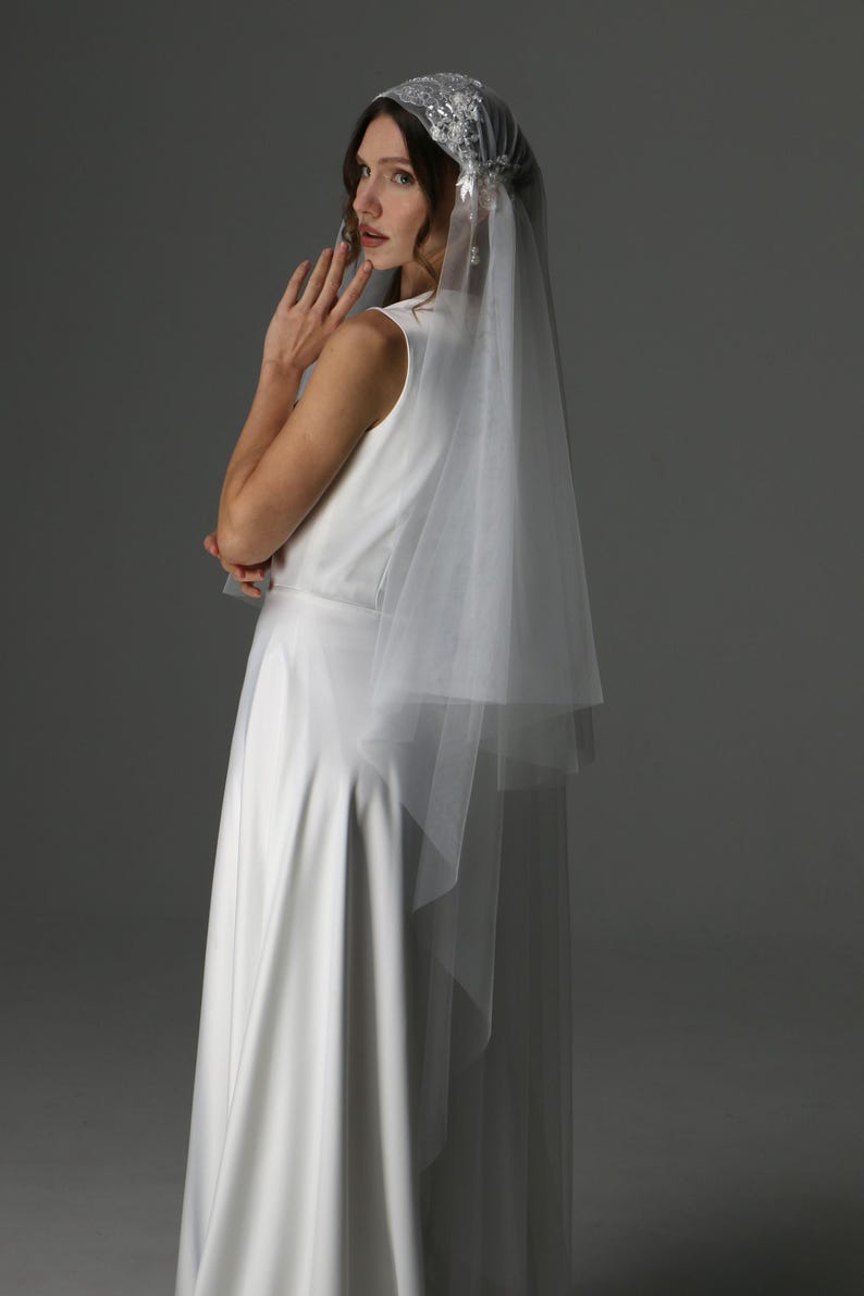 May include: A white bridal veil with a delicate, embellished headpiece. The veil is layered and falls gracefully. The bride is wearing a sleeveless white dress. The image is set against a neutral gray backdrop, highlighting the elegant details of the bridal wear.