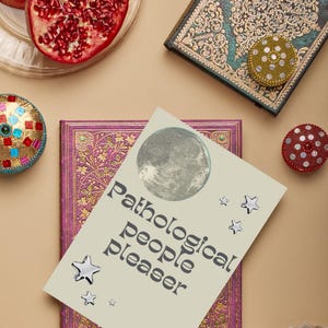 May include: An overhead shot features a journal with the text "Pathological people pleaser" and a moon graphic. Decorative books, a pomegranate, and embellished trinkets in various colors surround the journal. The scene is set on a neutral background.