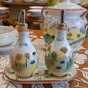 Può includere: Bottiglie per olio e aceto in ceramica con vassoio abbinato, con un design paesaggistico dipinto con alberi e colline. Le bottiglie hanno beccucci in metallo e tappi in sughero. Una ciotola da portata in ceramica abbinata e altre stoviglie in ceramica sono sullo sfondo.