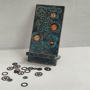 May include: A steampunk-themed phone stand with a distressed teal and bronze finish. The stand features clock faces and gears, with scattered metal gears at the base. The stand is designed to hold a phone or tablet.