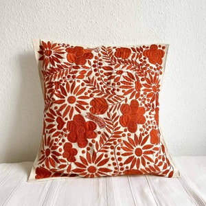 May include: A white pillow with a rust orange floral embroidery design. The pillow is made of a soft, natural fabric and features a variety of floral motifs, including flowers, leaves, and vines.