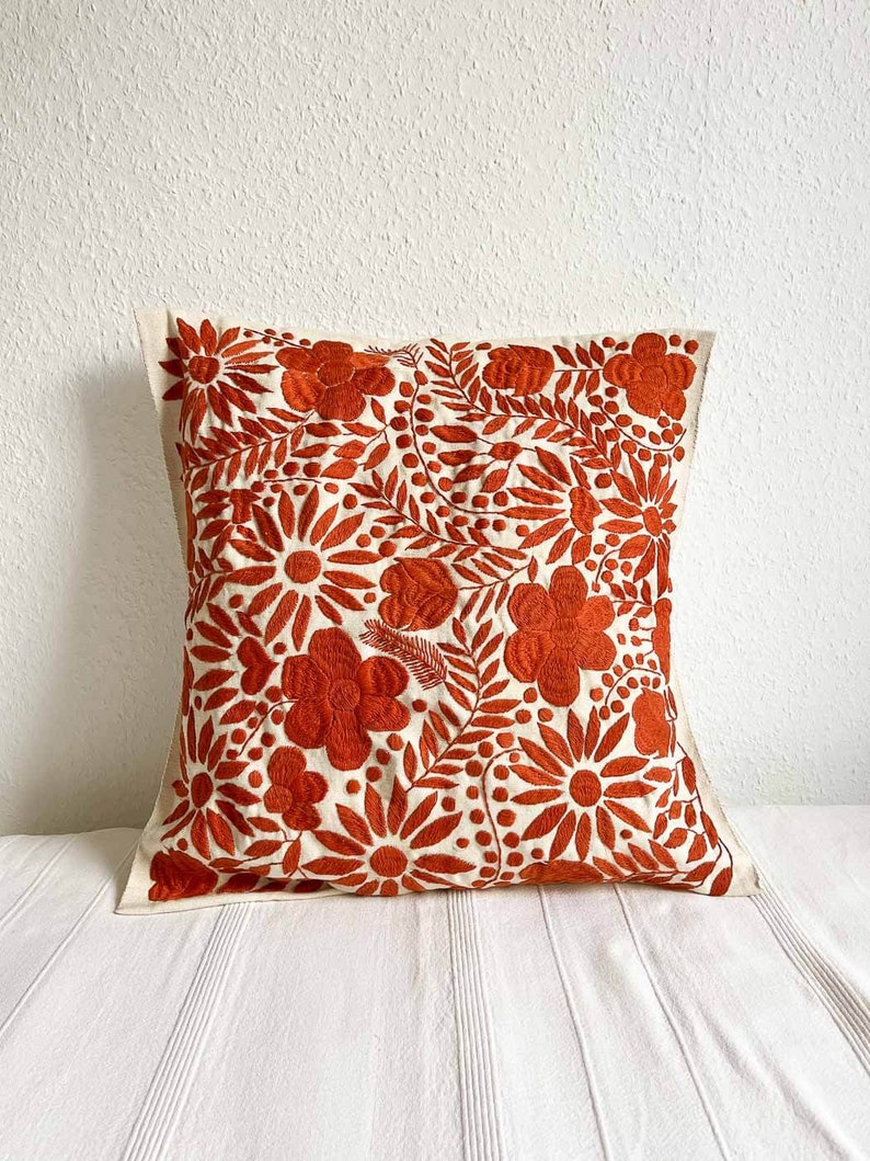 May include: A white pillow with a rust orange floral embroidery design. The pillow is made of a soft, natural fabric and features a variety of floral motifs, including flowers, leaves, and vines.