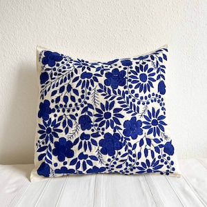 May include: A white pillow with a blue floral embroidery design. The pillow is made of a soft, textured fabric and features a variety of floral motifs, including leaves, flowers, and berries.