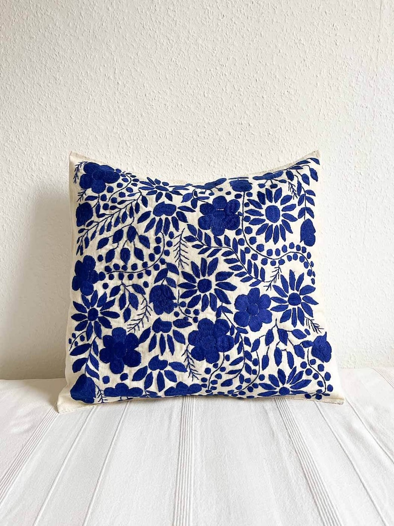 May include: A white pillow with a blue floral embroidery design. The pillow is made of a soft, textured fabric and features a variety of floral motifs, including leaves, flowers, and berries.