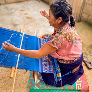 May include: A woman sits on a colorful mat and weaves a blue fabric on a wooden loom. She is wearing a pink blouse with floral embroidery and a dark blue skirt.