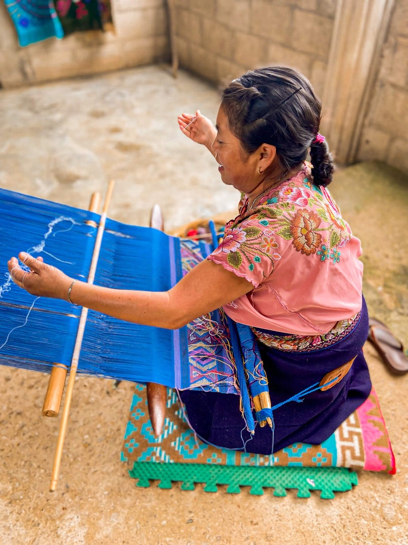 May include: A woman sits on a colorful mat and weaves a blue fabric on a wooden loom. She is wearing a pink blouse with floral embroidery and a dark blue skirt.