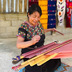 May include: A woman wearing a colorful embroidered blouse and black skirt sits on a woven rug and operates a wooden loom with red and pink threads.