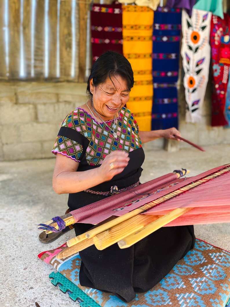 May include: A woman wearing a colorful embroidered blouse and black skirt sits on a woven rug and operates a wooden loom with red and pink threads.