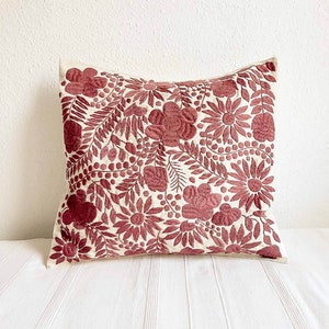 May include: A white pillow with a red floral embroidery design. The pillow is made of a soft, natural fabric and features a variety of floral motifs, including flowers, leaves, and berries.