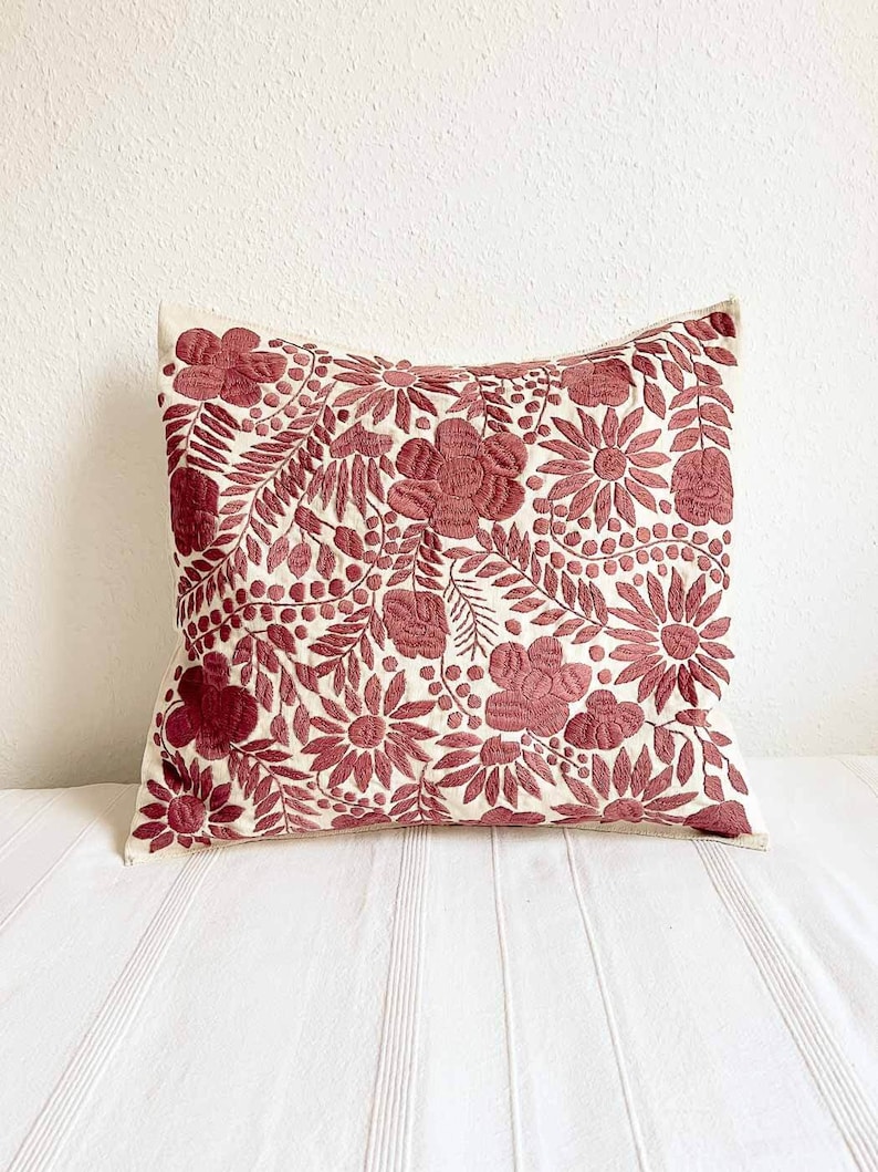 May include: A white pillow with a red floral embroidery design. The pillow is made of a soft, natural fabric and features a variety of floral motifs, including flowers, leaves, and berries.