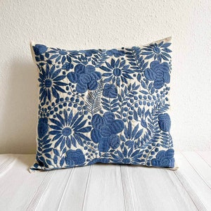 May include: A blue and white embroidered pillow with a floral pattern. The pillow is made of a soft, natural fabric and has a square shape.