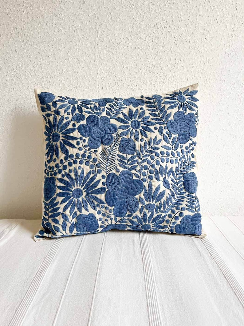 May include: A blue and white embroidered pillow with a floral pattern. The pillow is made of a soft, natural fabric and has a square shape.